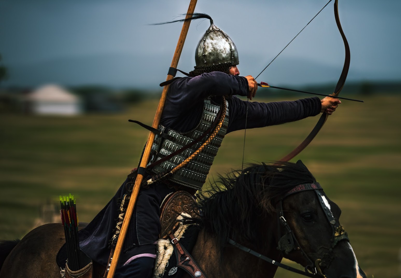 A mounted archer seen from behind, drawing a recurve bow, helmet plume flying