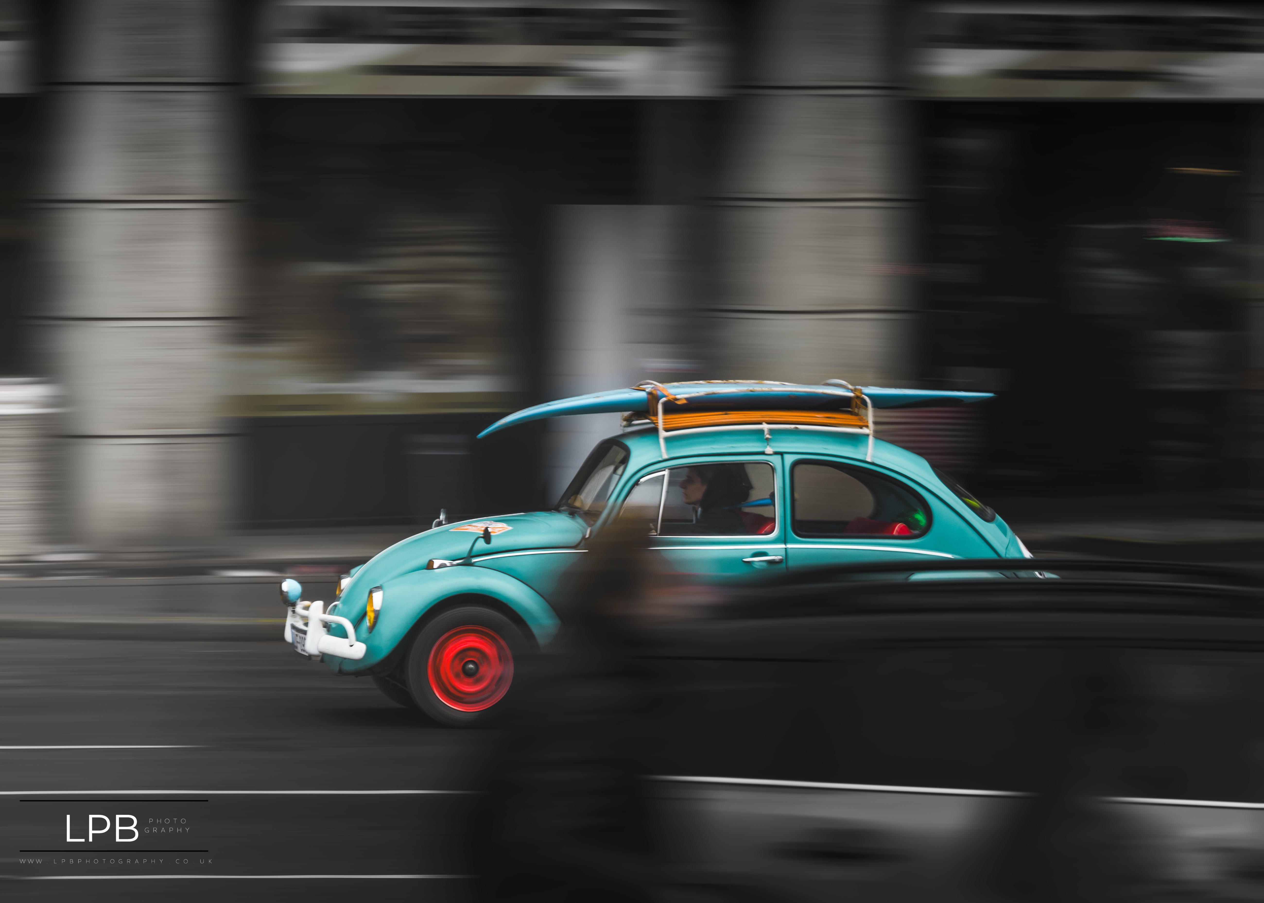 A teal VW Beetle with a surfboard, sharp against a fully motion-blurred street, red wheel glowing