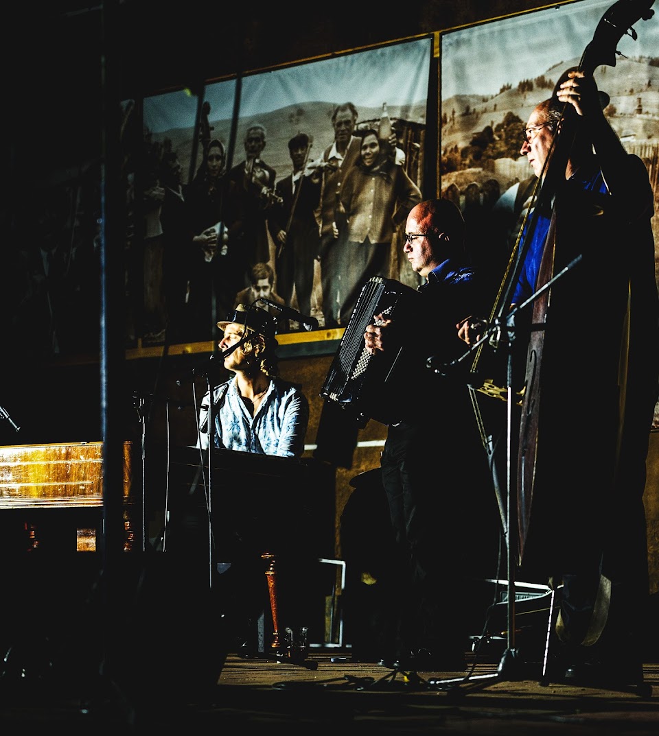 A trio at the keys, accordion and double bass, lit against a large projected archival photograph of village musicians