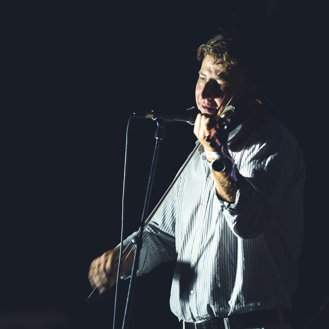 A violinist at the microphone in dramatic side-light, a keys player beside